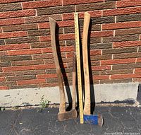 Two rustic axes standing upright next to a ruler against a brick wall. One axe has a brown handle; the other has a lighter handle and blue painted axe head.