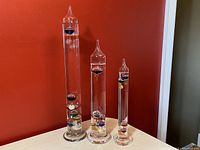 Full view of the set of three graduated height Galileo thermometers on white surface against red wall.