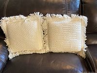 Two vintage cream white crochet throw pillows with fringe edging resting on a dark leather sofa. One pillow has a diamond-like textured crochet pattern; the other has a checkerboard-like textured crochet pattern.