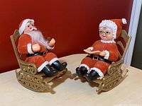 Front view of Santa Claus and Mrs. Claus flocked figures seated on plastic rocking chairs against a red background.