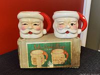 Two ceramic Santa Claus mugs with winking faces sitting on top of their original box. The box label reads 'Jolly Santa Mugs 2-to-set' with images of the mugs. The mugs have red hat handles and visible paint wear.