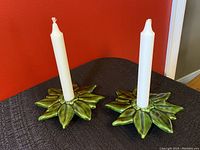 Two green ceramic poinsettia-shaped candle holders with white tall tapered candles inserted, set on a black surface with red wall background.