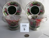Two Royal Albert Country Fayre Somerset tea cups wrapped in original plastic bags placed in front of matching saucers with green center, floral and fruit design, and scalloped gold-trimmed edges.