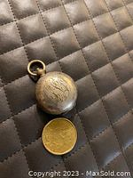 Exterior view of round silver coin holder with engraved initials, closed, beside a gold coin for size comparison.
