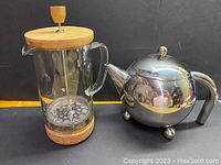Photo showing both the French Press with glass body, solid wood lid, cork base, and the stainless steel spherical tea pot with three rounded legs and black handle.