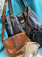 Three leather bags (black large handbag, smaller black bag, brown crossbody) and a light brown/cream wool scarf with tags displayed together