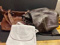 Three leather bags displayed: large dark brown Roots bag, medium brown tote bag, small light colored flap bag