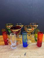 Lot of mixed vintage glassware with six ornate small goblet-style wine glasses and six colorful shot glasses with handles arranged on a wood surface against a black background.