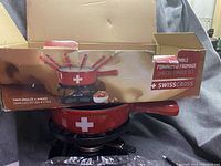Photo showing the entire fondue set with pot in stand and red-handled forks inside. Box with Swiss Cross branding and product details included.