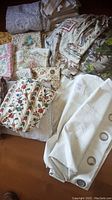 Assorted drapes, curtains, and fabric window coverings laid on a flat surface showing patterns, textures, and some white curtains with metal grommets.