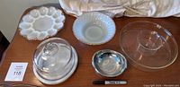 Six pieces of serveware on table including a glass dome with marble base, a white milk glass devilled egg platter with gold scalloped edge, a white fluted glass bowl, a clear glass Anchor Hocking bowl with gold trim, a stainless steel Sambonet Twist oval serving dish, and a marker for scale.