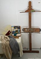 Photo showing full view of adjustable wooden easel next to a table with artist supplies and smocks.