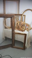 Full view of multiple ornate and gold leaf picture frames stacked and leaning against wall and table, shown in different sizes and shapes, including oval and rectangular.