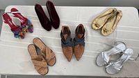 Overview of six pairs of women's shoes on white surface including heels, flats, sandals in various colors and materials.