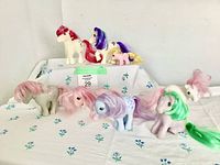 All 11 My Little Pony dolls arranged on a table covered with floral cloth. Various colors of manes and tails visible. Dolls have painted eyes and flank symbols, some are unicorns.