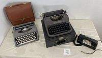 Overview photo showing both typewriters with their cases and rotary dial telephone on a white table.