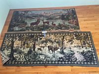 Photo of two woven fabric wall hangings laid on wooden floor, one longer with elk and forest scene, the other shorter with deer and lake scene.