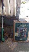 Photo showing the boxed 4 tier mini greenhouse with visible design and brand labeling, placed on the floor alongside the green Weed Eater trimmer edger handle and shaft.