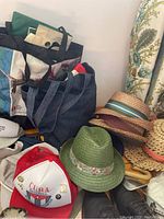 Photo of several hats stacked beside reusable bags, including green fedora with floral band, beige straw hats, and a red and white baseball cap.