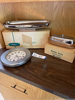 Photo of vintage Milbern chromium bread tray, butter dish with original boxes, and quartz wall clock on wooden shelf.