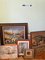 Photo showing multiple framed vintage paintings depicting churches and rural landscapes assembled on floor against wall