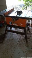 Side view of orange and black table saw with blade and fence on top, metal legs, and adjustment wheel on side.