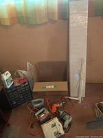 Overall view of the power tools, blades, and shelf packed together on the ground by the wall, some in cardboard box, shelf leaning against the wall with brackets tied