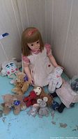 3-foot vintage plastic doll seated against corner wall with several teddy bears and cloth dolls surrounding it on blue floor