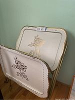 Stacked set of four vintage metal TV trays with rose patterns visible on surfaces and gold scalloped edges. Showing slight wear from use.