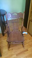 Front view of vintage wooden rocking chair showing carved backrest, spindles, seat and armrests on hardwood floor.