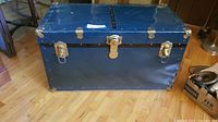 Front view of the vintage blue metal steamer trunk with metal handles and latch, sitting on hardwood floor.
