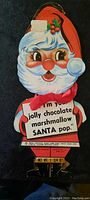 Front view of the cardboard Santa Claus advertising card showing Santa's face, hat with holly decoration, and jolly expression with text promoting chocolate marshmallow Santa pop.