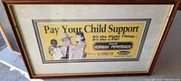 Framed rectangular vintage political sign with yellow background, messenger text about child support, image of sheriff and children. Shows wood frame and glass cover.