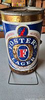Front view showing cylindrical tin grill painted with Foster's Lager branding and colorful graphics with signs of surface rust and wear.