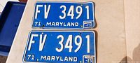 Two blue Maryland license plates from 1971 with white embossed letters and numbers 'FV 3491', stickers intact, minor bends visible