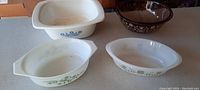 Four vintage bakeware pieces shown on a countertop including three white Corningware dishes with floral patterns and one brown Pyrex smoked glass bowl with floral geometric design.
