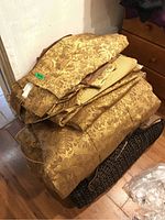 Stack of folded gold damask patterned comforter, pillow shams, and other matching fabric pieces on a floor next to a dresser.