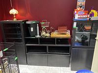 Full view of three black manufactured wood cabinets positioned side by side. Left cabinet has 2 solid doors, middle cabinet has 2 drawers and open shelving topped with glass surface, right cabinet has 2 glass doors. Various items placed on top and shelves, cabinets closed.