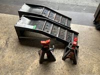 Two metal car ramps laid side by side with two black Motomaster axle stands with red locking mechanisms in front, on floor.