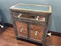 Front view of the console table showing wood veneer frame, mirrored drawer front with two clear glass knobs, two mirrored cabinet doors with x-pattern overlays and glass knobs, scratches and crack on drawer front visible.