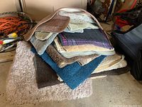 Stack of assorted floor mats in various colors and textures on a concrete floor with other items