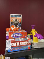 Stack of three boxed board games including Battleship, Operation Shrek, and The Best of TV & Movies with a toy clownfish figure on top set against a red wall.