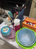 Overview of kitchenware on table including plastic bowls, storage containers with lids, thermos, mugs, utensils in fabric holder, and serving platters