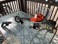 Photo of the Black & Decker leaf blower model CS100 in orange and black, a Yardworks electric trimmer with green guard and metal shaft, charger for the leaf blower, all arranged on a glass outdoor table.
