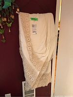 Cream linen blanket with wide crochet lace trim draped over a white hanger against a dark red wall.