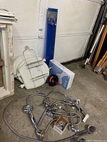 Wide shot showing three white toilet seats stacked, blue boxed bidet attachment, plumbing snake, and various hoses and shower heads on concrete floor.