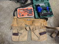 Overview of Power Fist nailer/stapler in green case, orange case with hand tools, and canvas tool pouch with belt laid out on concrete floor.