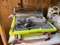 Photo of the Ryobi tabletop wet tile saw showing green base, metal tabletop, circular blade with cover, miter guide, and power cord.