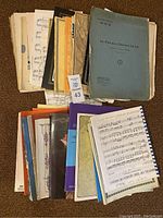 Top view of a large assortment of worn sheet music including several books and loose pages in various colors, stacked and fanned out on a carpeted floor.