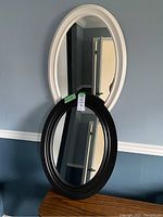 Both black and white oval mirrors stacked against a wall, showing overall shape, frame color and size relative to surroundings.
